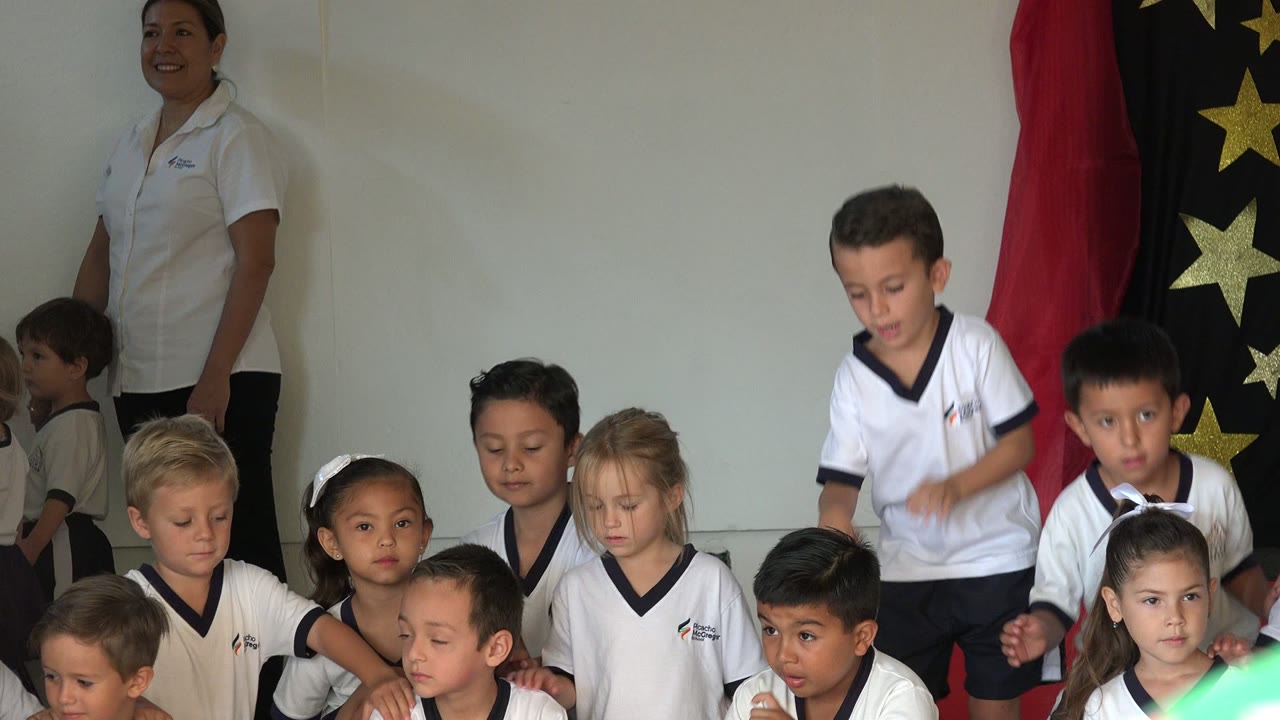 A standing boy looks downward among seated classmates in San Jose del Cabo, Mexico, with the teacher and star-patterned black fabric behind the group.