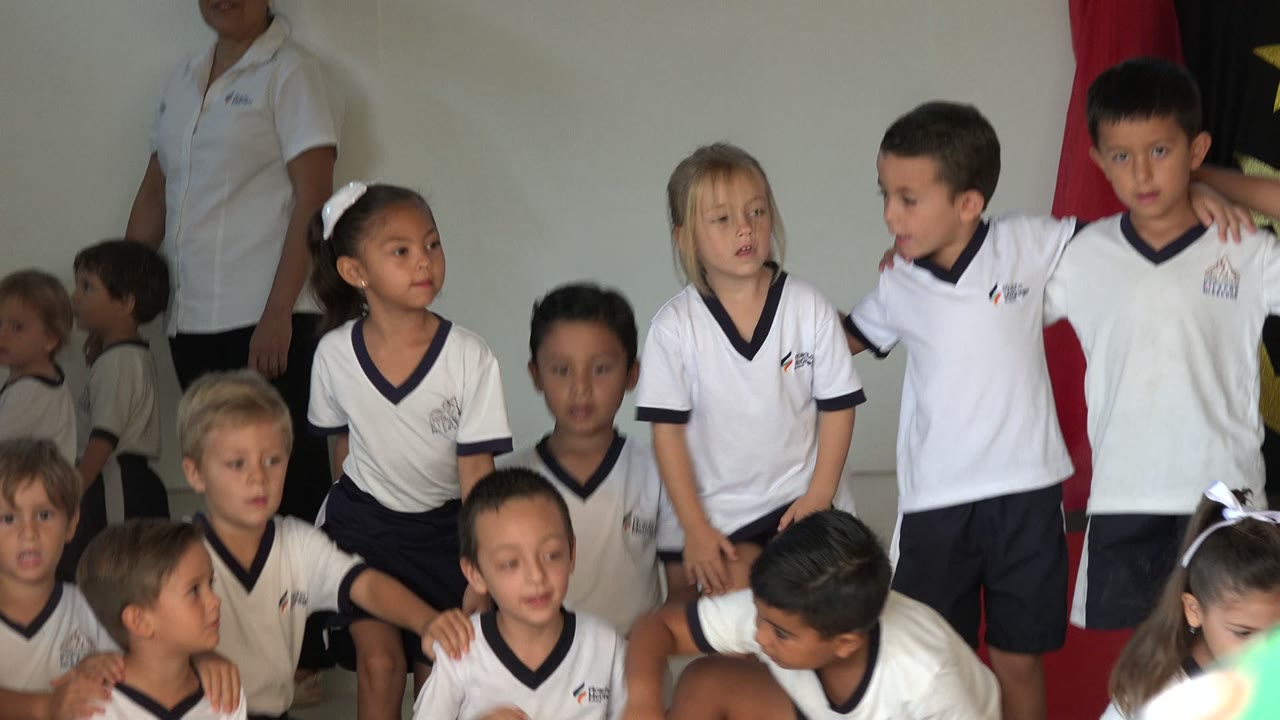 Children in white uniforms stand together with an adult partly visible behind them.
