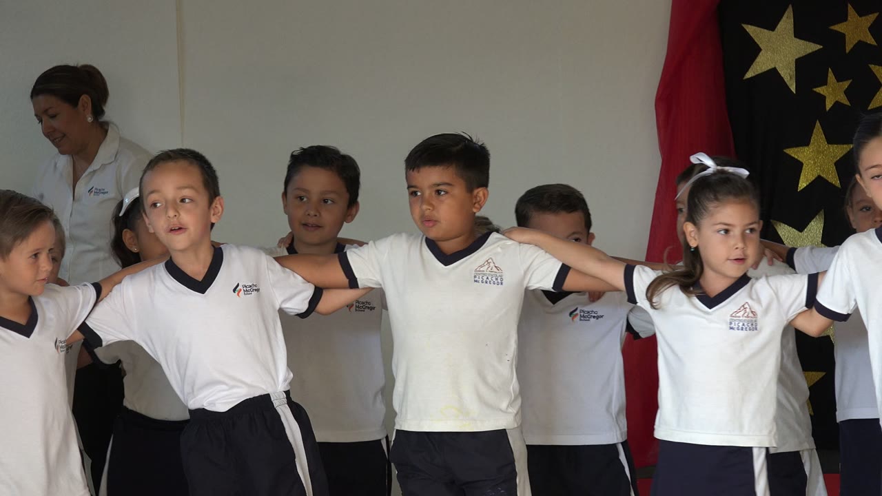 Children in white school uniforms stand with arms out during a group performance.