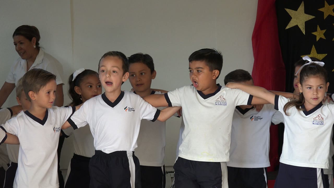 Several children in white shirts and dark shorts continue singing or performing together in San Jose del Cabo, Mexico, with a black and gold star backdrop behind them.