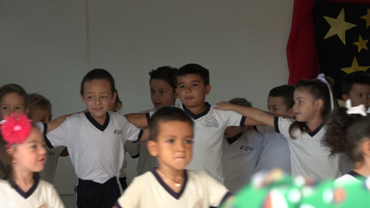 Children in white shirts stand with linked arms indoors.