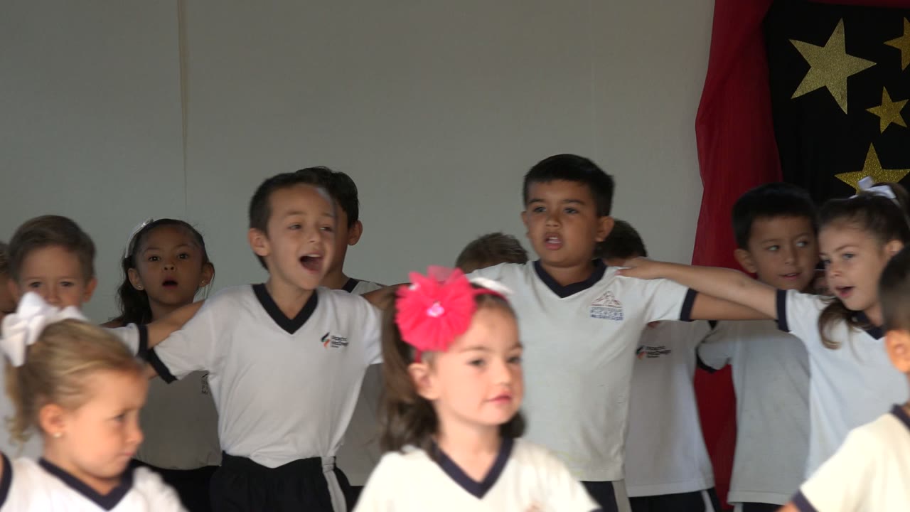 A boy near center sings with his mouth open as children in matching white shirts link arms behind foreground girls in San Jose del Cabo, Mexico.
