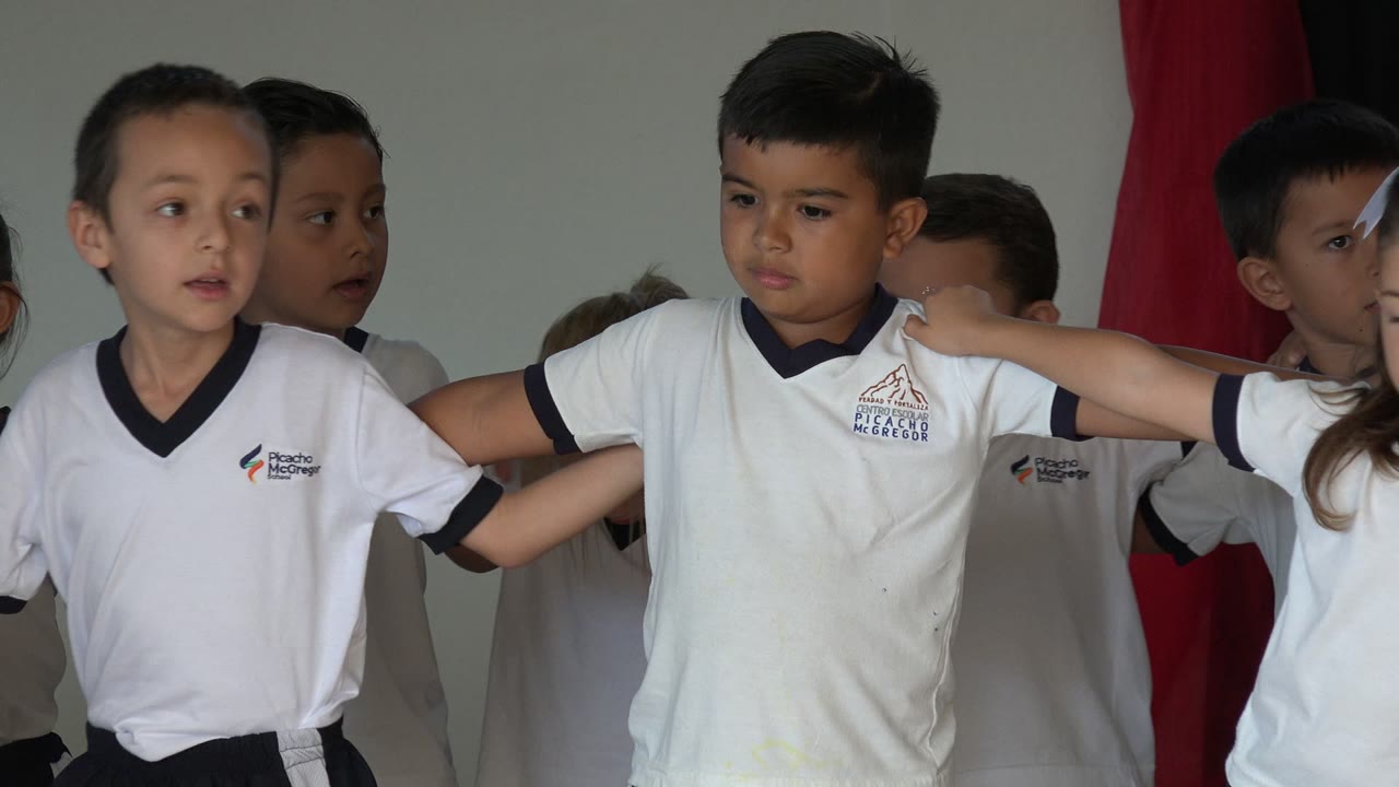 Children in white shirts stand linked arm in arm on an indoor stage.