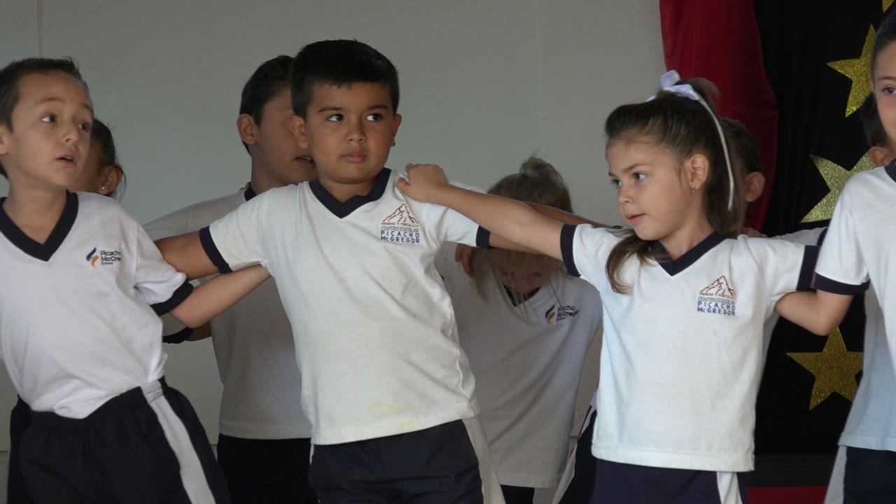 A row of young children in San Jose del Cabo remain linked across the stage, with one girl in a white hair bow looking toward a boy beside her.