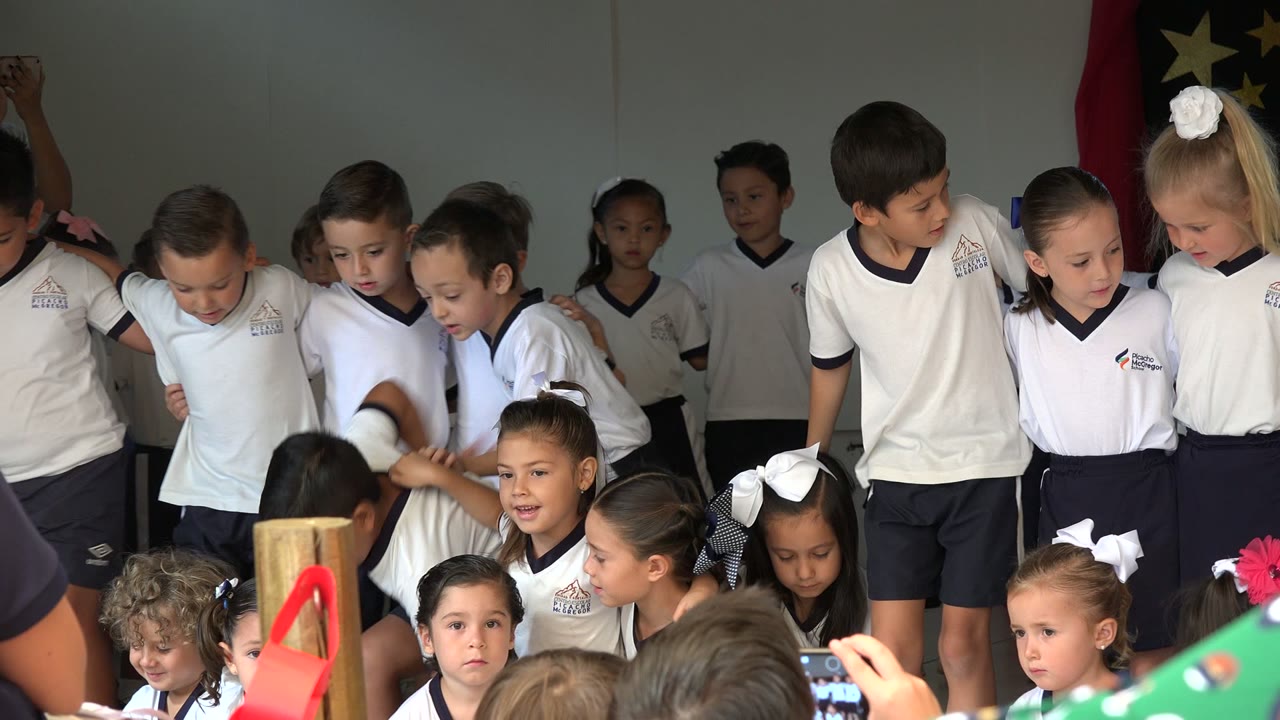 A line of uniformed children in San Jose Del Cabo, Mexico shifts in place, with a tall boy near center looking down and younger children seated below the stage line.