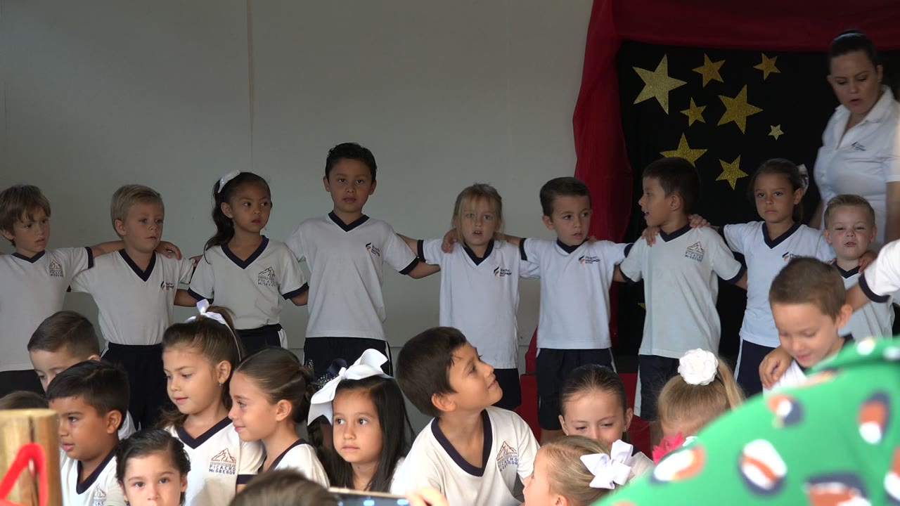 Children in San Jose del Cabo stand arm in arm across a stage area while an adult in a white shirt stands near the back and seated children fill the foreground.