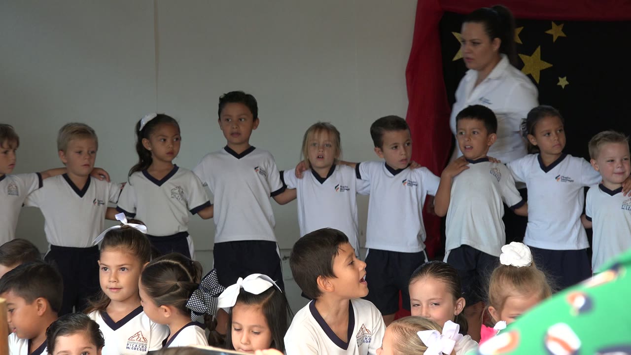 A line of children in white shirts stands arm in arm in San Jose del Cabo, with several children looking toward the room and a black backdrop with gold stars behind them.