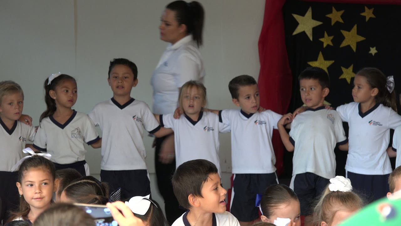 Children in matching white shirts stand together while an adult passes behind them.