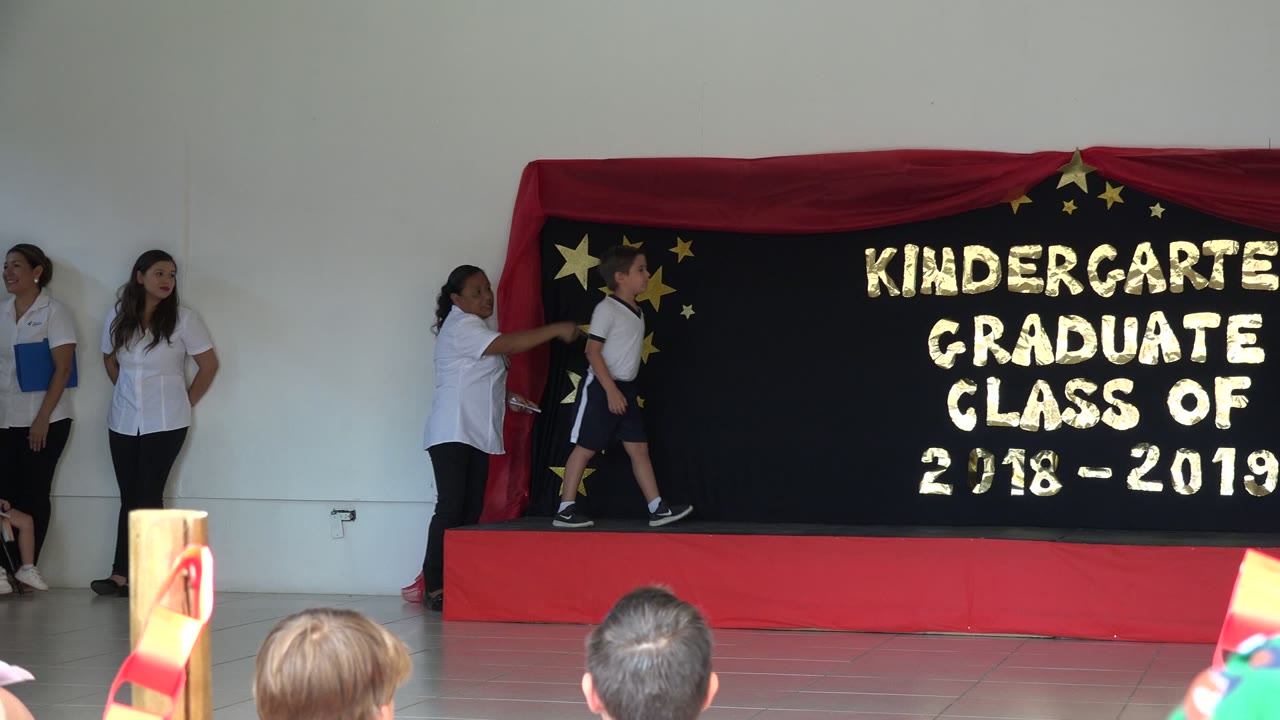 Boy walking on a red stage beside women in white shirts at a kindergarten graduation.