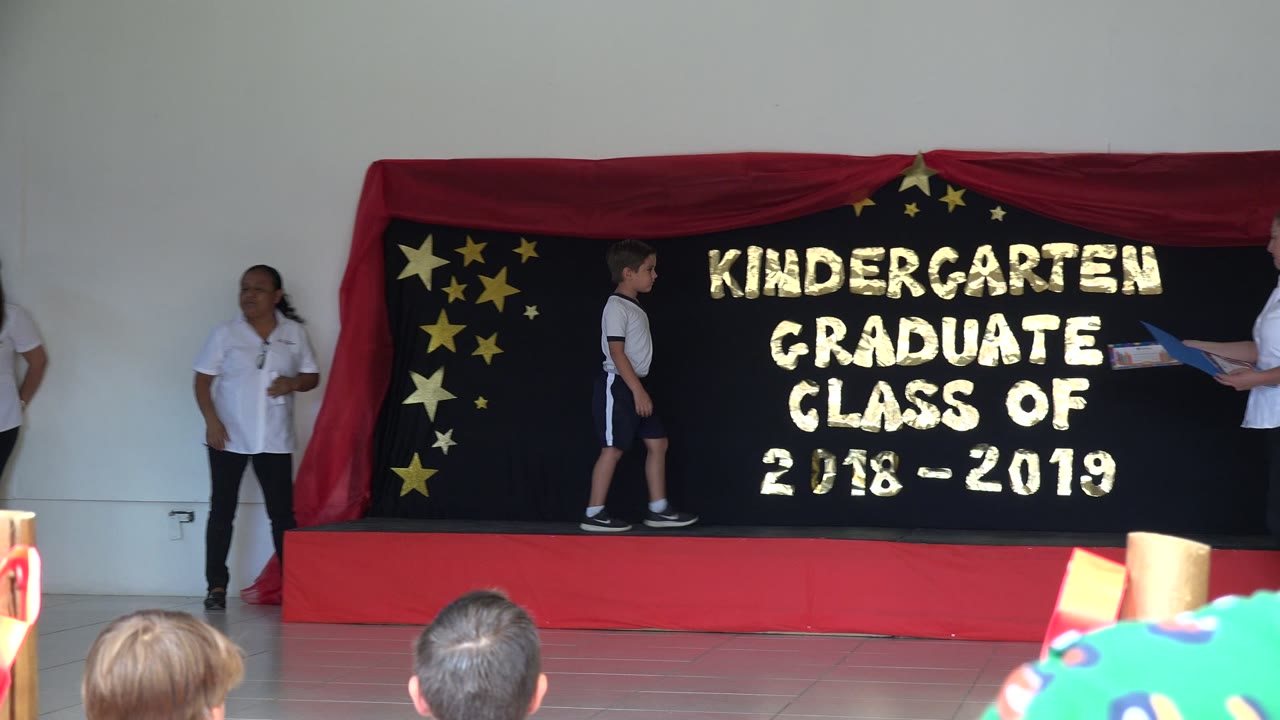 The boy pauses near center stage in front of a black backdrop reading kindergarten graduate class of 2018-2019, with adults standing at both sides and audience heads below.
