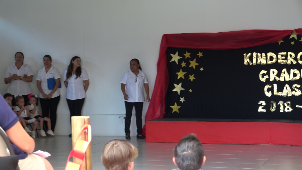 School stage with teachers, seated children, audience heads, and a kindergarten class backdrop.