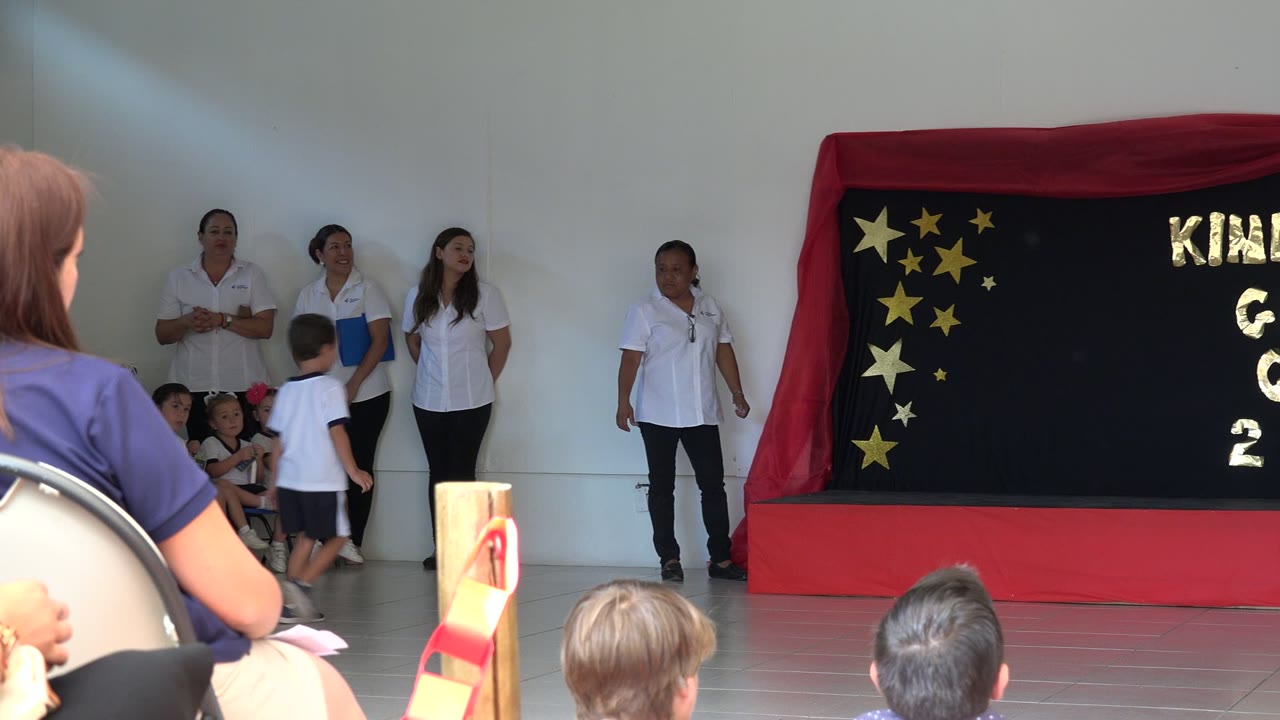 A boy walks across the stage area in San Jose del Cabo, Mexico, while three women in white tops stand near seated children and a decorated kindergarten backdrop.