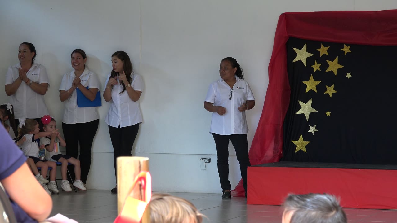 Women in white shirts stand near seated children and a star-covered stage backdrop.