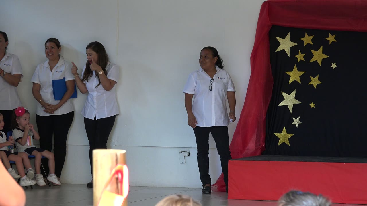 In San Jose del Cabo, Mexico, a woman in a white shirt stands near the red stage platform as children sit at the edge of the star-decorated backdrop.