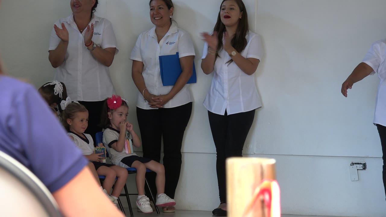 In San Jose del Cabo, Mexico, adult women in white shirts stand against a plain indoor wall while two young girls sit nearby and a foreground object partially blocks the lower frame.
