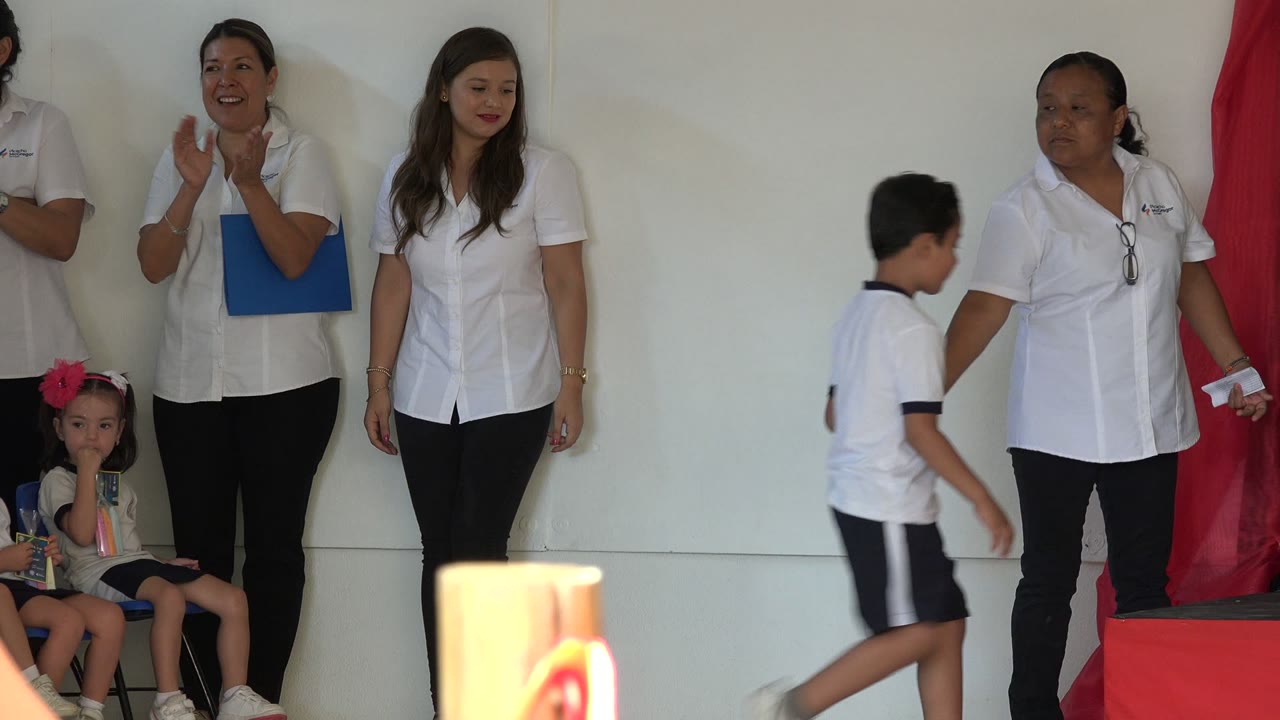 Women in white shirts stand near seated children during an indoor event.