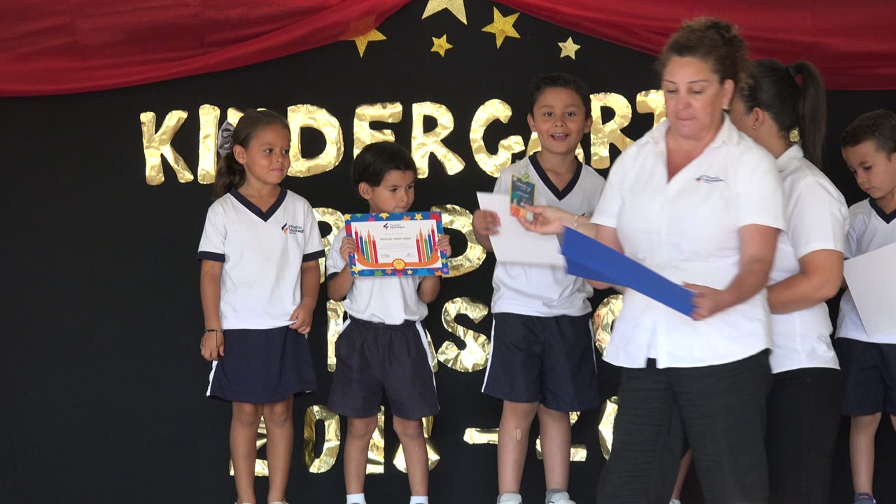In San Jose Del Cabo, Mexico, children in uniforms stand near an adult holding a blue folder as a decorated certificate or craft is displayed against the kindergarten backdrop.