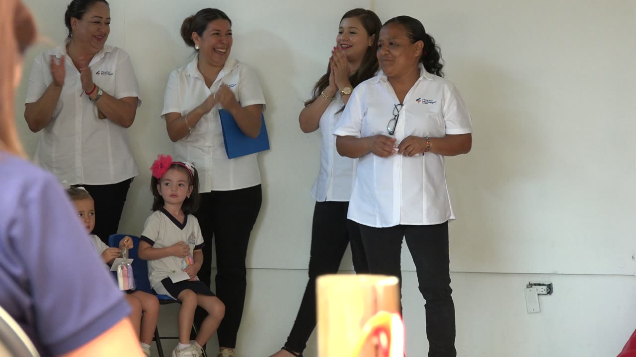 Women in white shirts stand by a wall with seated children below.