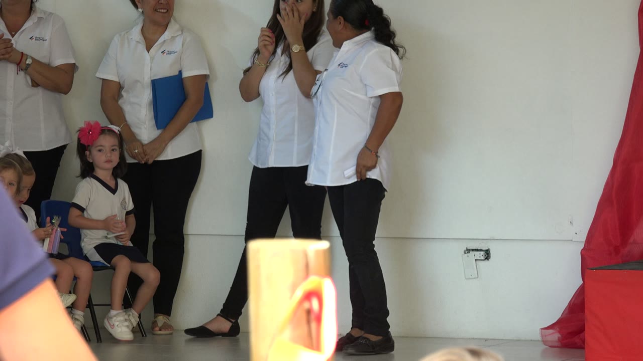 Adults in white shirts stand near seated children during an indoor ceremony.