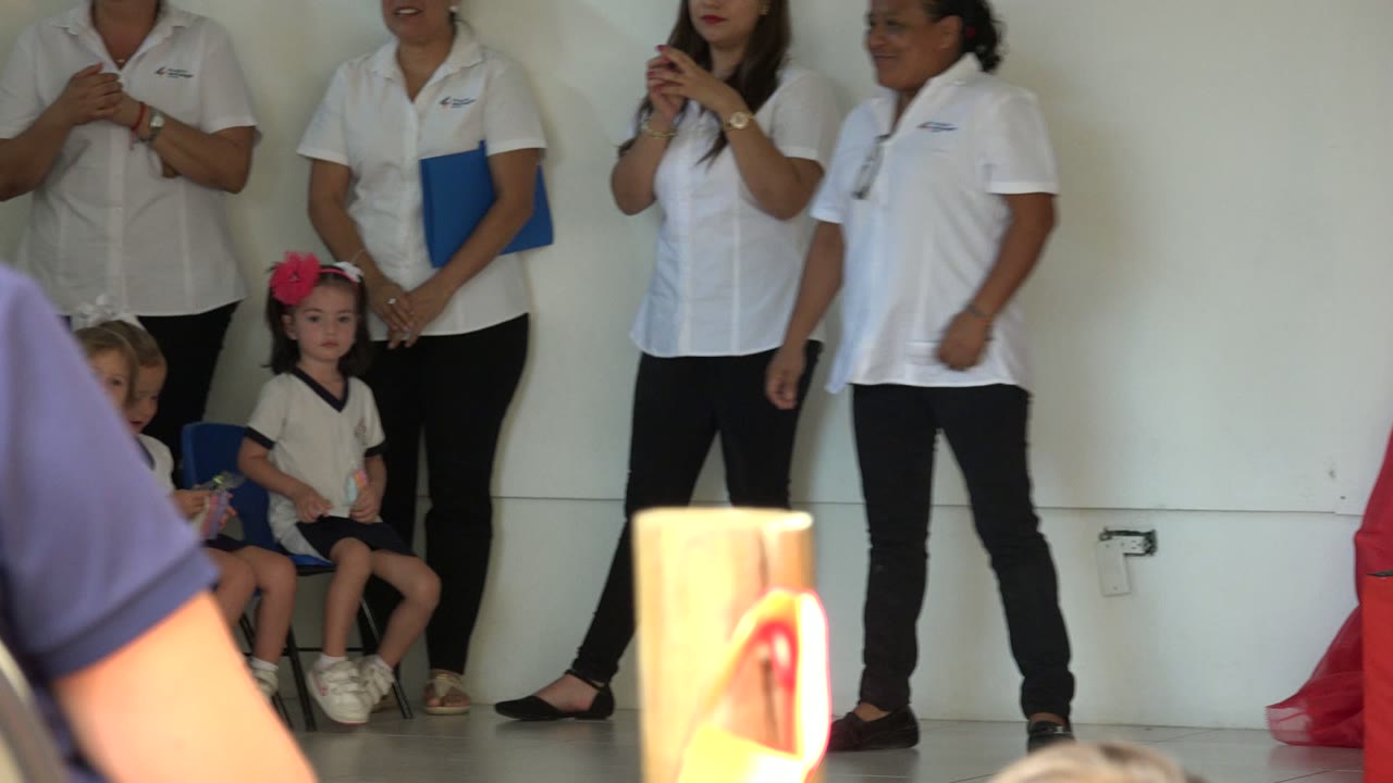 Women in white shirts clap near seated children in San Jose del Cabo, with a child wearing a pink hair bow facing the camera and a foreground audience member at left.