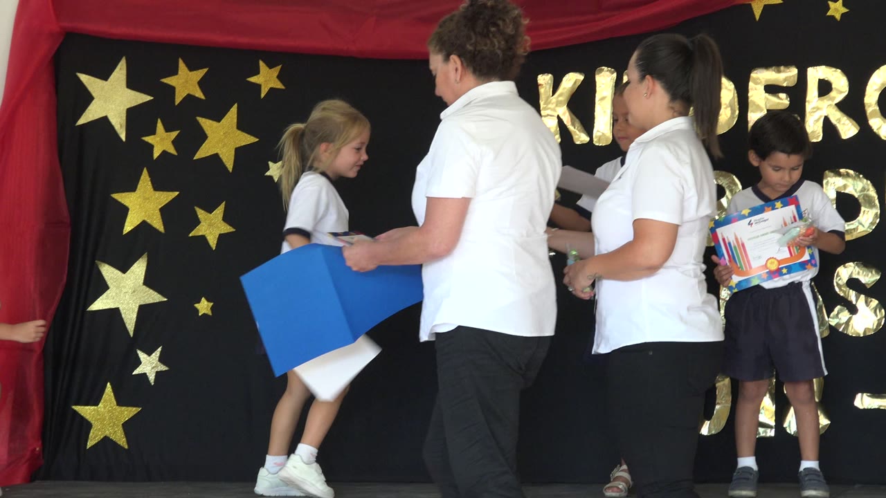 Woman in a white shirt hands papers to children on a decorated stage.