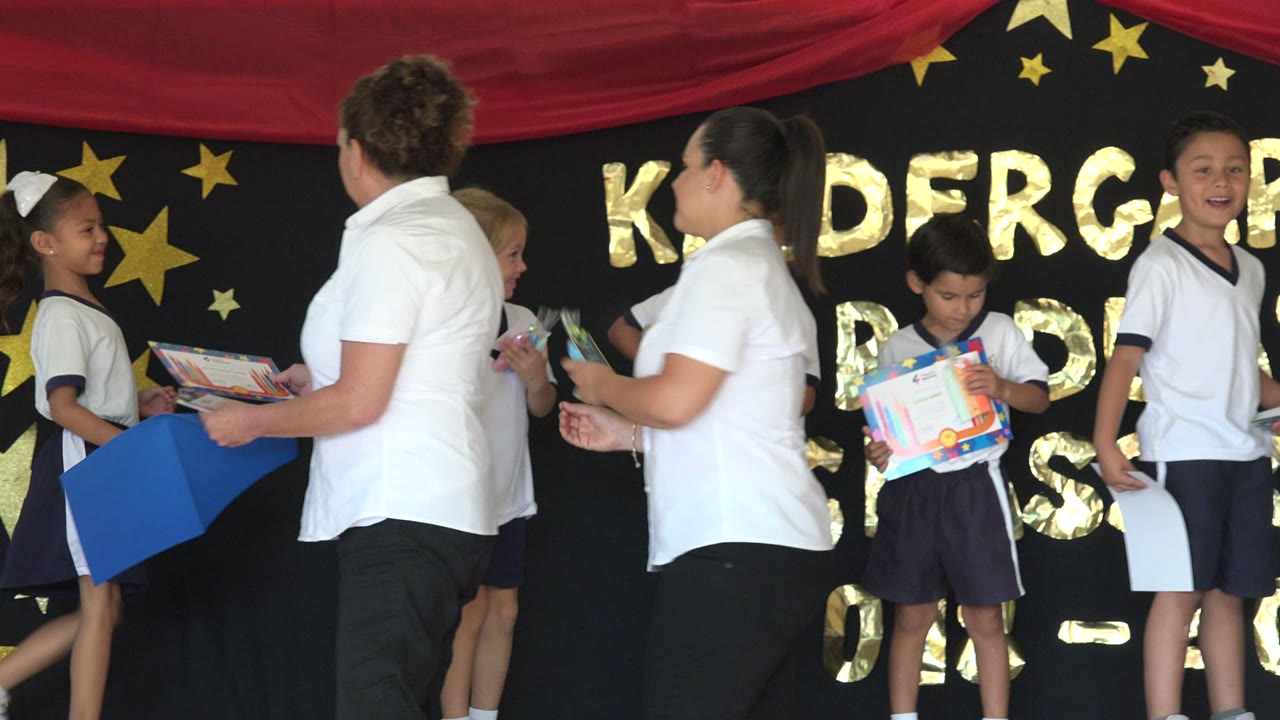 In San Jose del Cabo, Mexico, several children in white uniforms hold colorful certificates while two women in white shirts stand nearby on the decorated stage.