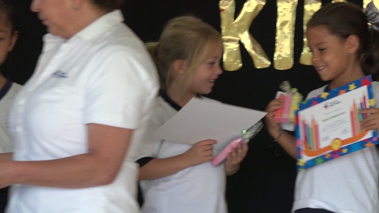 At 0:01 in San Jose Del Cabo, Mexico, a girl holds a colorful certificate and small wrapped gift while adults in white shirts partially block the foreground.