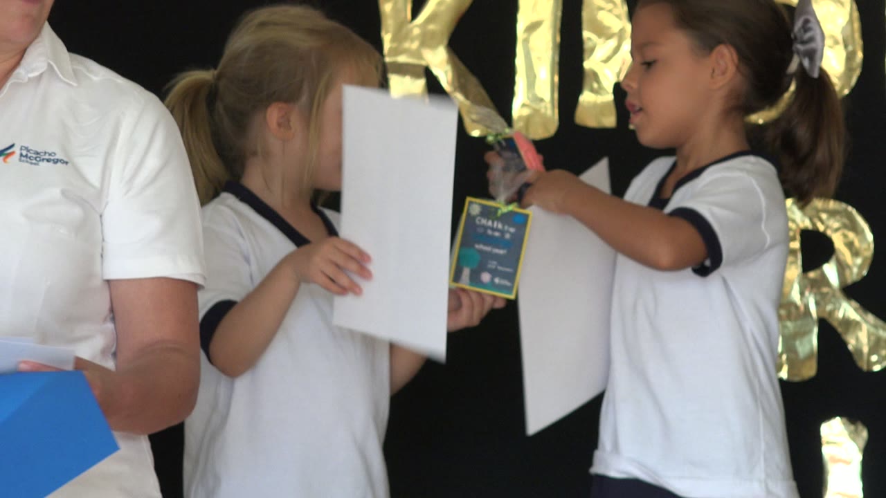 Children in white school shirts hold papers during an assembly.