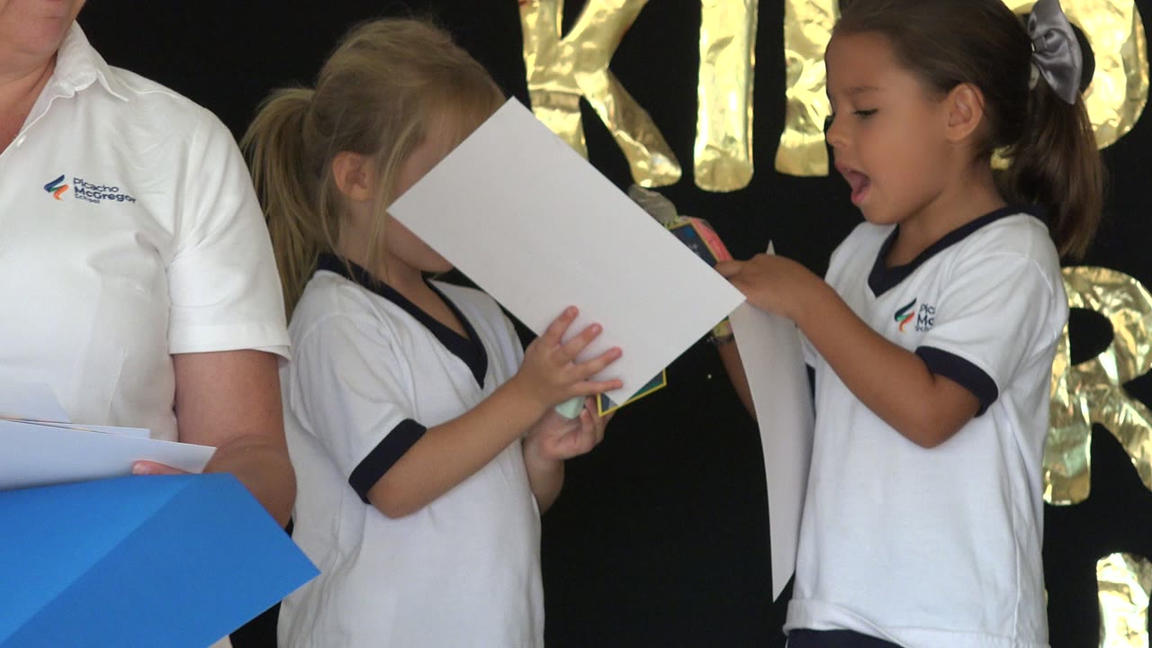 A San Jose Del Cabo assembly moment shows one child partly hidden behind a sheet of paper while another child holds a small item near a black curtain with gold decorations.