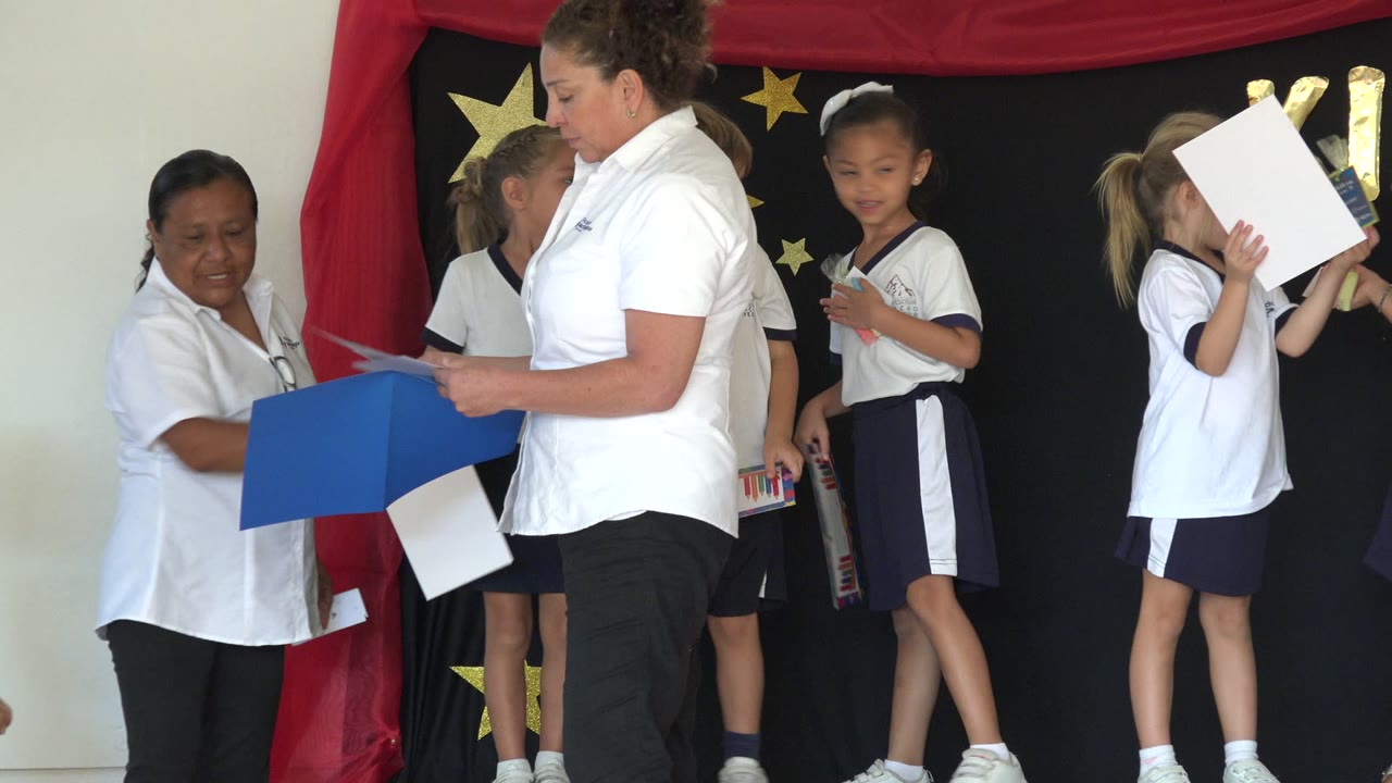 Teachers hand papers near children standing on a decorated kindergarten stage.