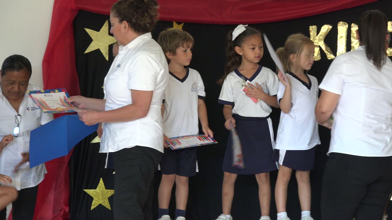 Children in white and navy uniforms stand in a row in San Jose del Cabo, Mexico, as one teacher holds a blue folder and another teacher crouches near certificates.