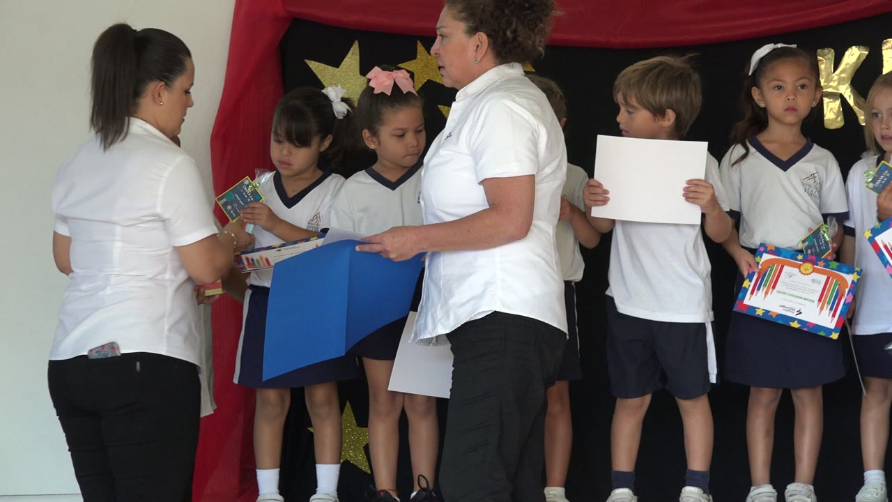 Teachers organize children on a small decorated school stage.