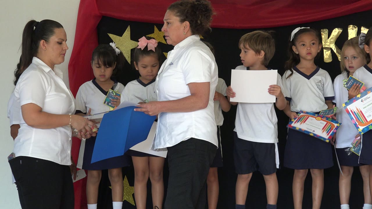 Two teachers stand near the front of the stage as children in uniforms hold papers and certificates against a red and black decorated backdrop in San Jose del Cabo, Mexico.