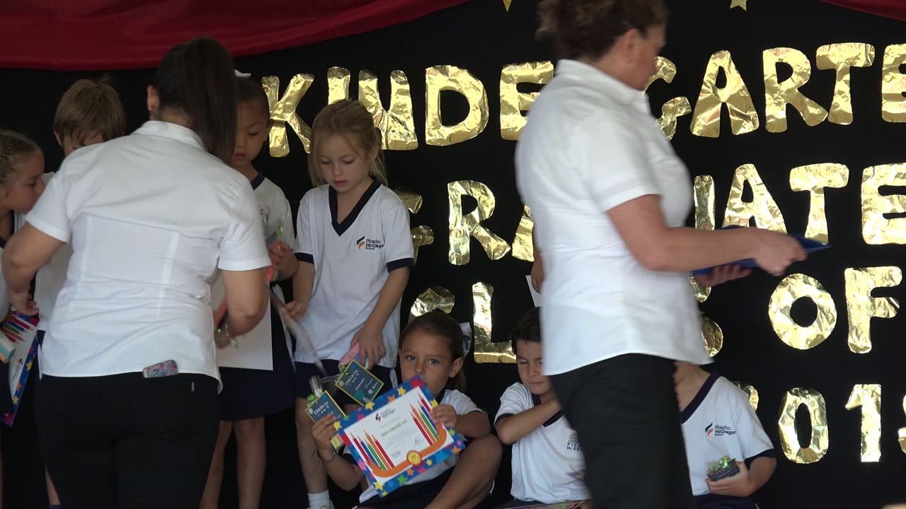 At 0:01 in San Jose del Cabo, Mexico, the teacher is seen from behind as several children sit with certificates and others stand near the gold-lettered backdrop.