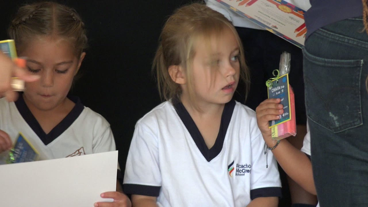 A young girl in a white school shirt looks down at papers while an adult in jeans stands close in the foreground in San Jose del Cabo, Mexico.