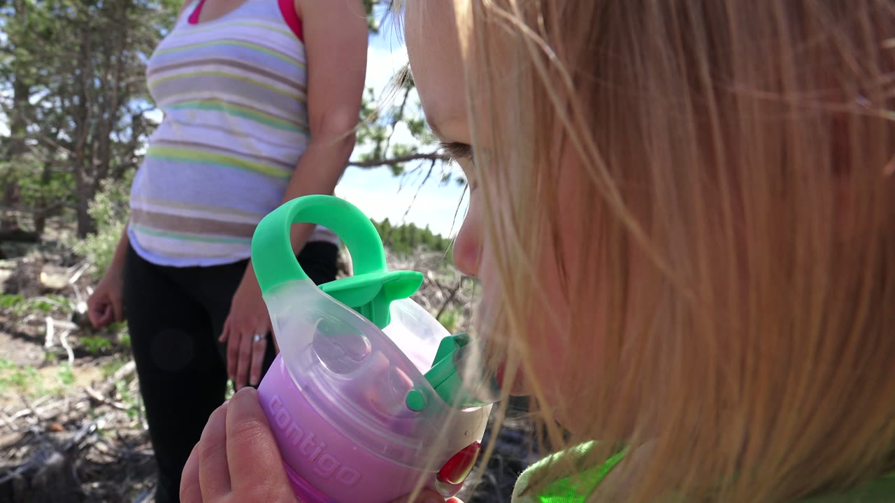 In Westminster, Colorado, a blond toddler is seen from behind and the side holding a purple and green sippy cup while an adult stands in the sunlit wooded background.