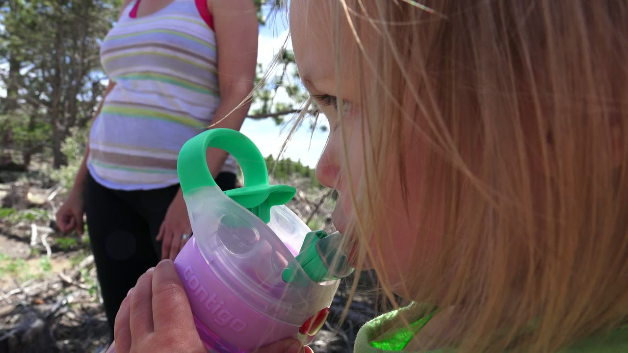 In Westminster, Colorado, the toddler turns more into profile as the purple and green sippy cup is held near their mouth, with the adult still cropped behind them.
