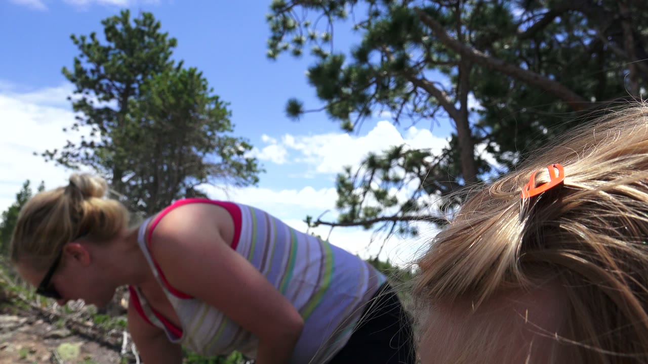 In Westminster, Colorado, the woman bends forward in sunglasses while the child's blond hair crosses the foreground against pine trees and blue sky.