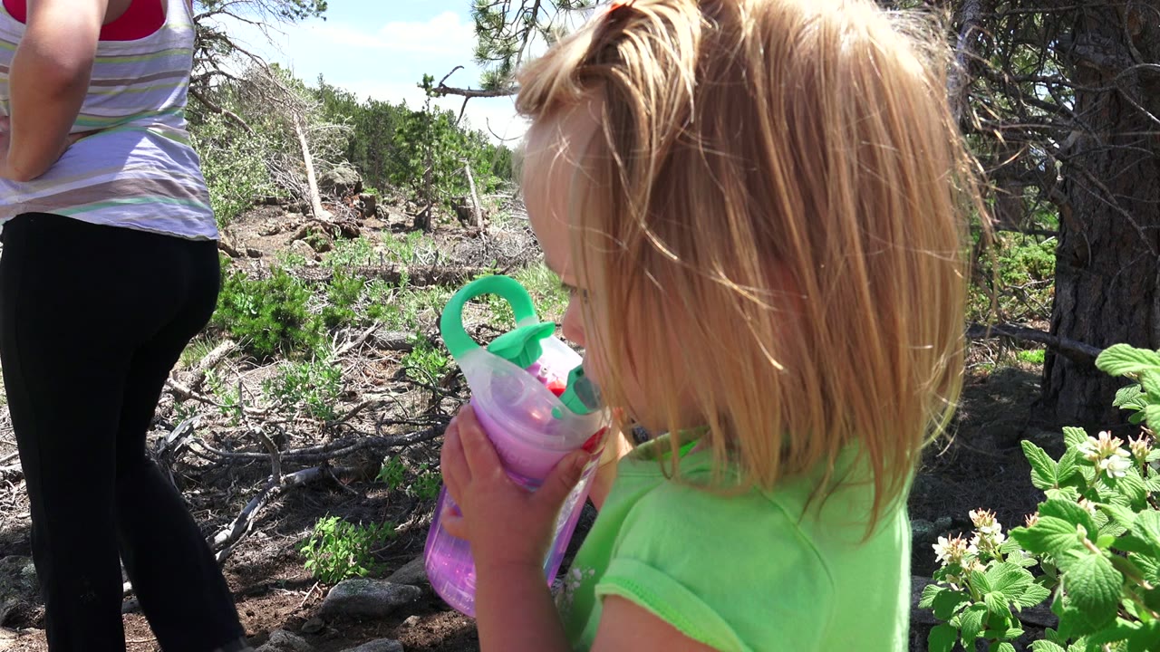 At 0:01 in Westminster, Colorado, the child remains in close foreground profile with the drink cup raised, while the adult is cropped at the torso in the sunlit brush.