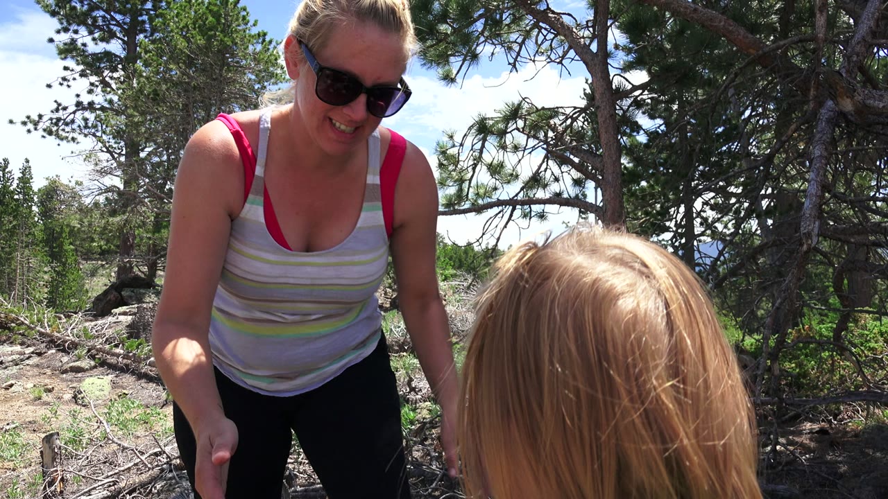 Woman in sunglasses leans toward a blonde child outdoors among trees.