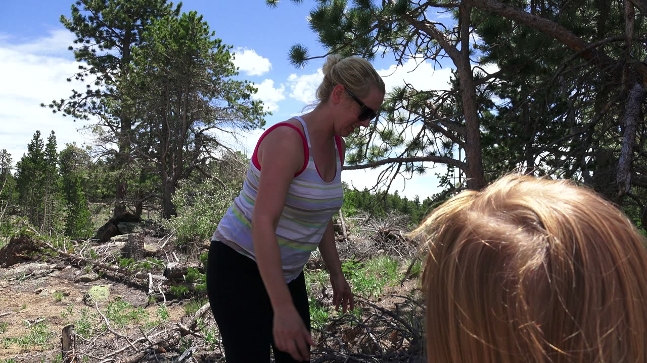In Westminster, Colorado, the woman stands slightly back from the blonde child in the foreground, surrounded by sunlit brush, logs, and pine trees.