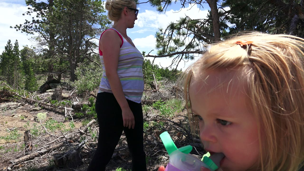At 0:01 in Westminster, Colorado, the young girl holds a purple cup with a green lid to her mouth while the adult woman stands in profile behind her.
