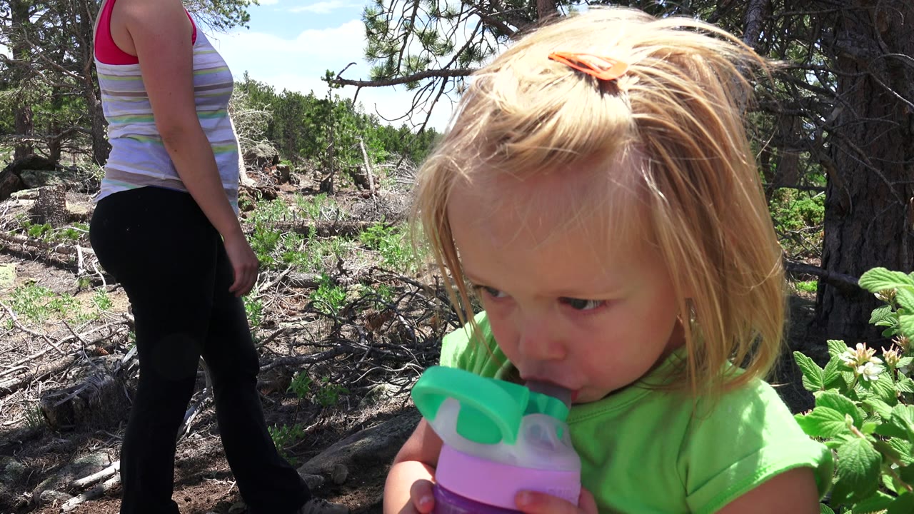 Toddler in green shirt drinks from a sippy cup beside an adult outdoors.