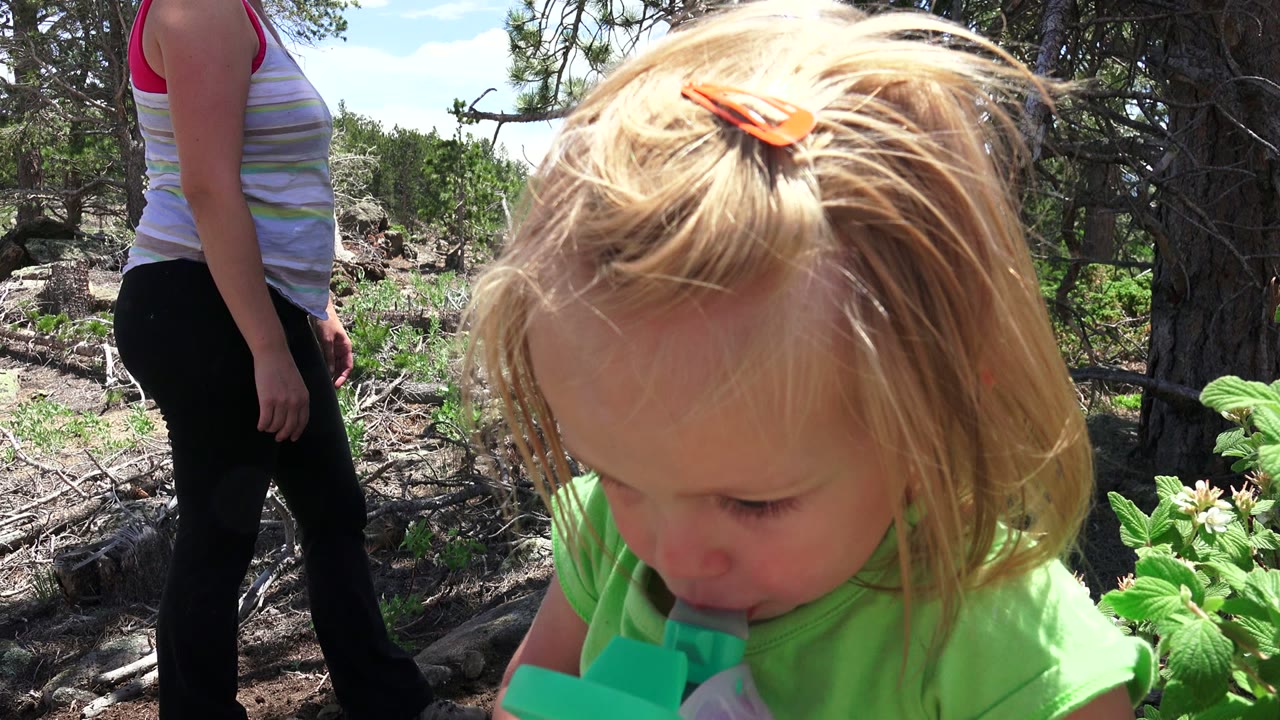 In Westminster, Colorado, the toddler remains close to camera with the sippy cup at their mouth, while the adult woman's torso and legs appear in the background near evergreen trees.