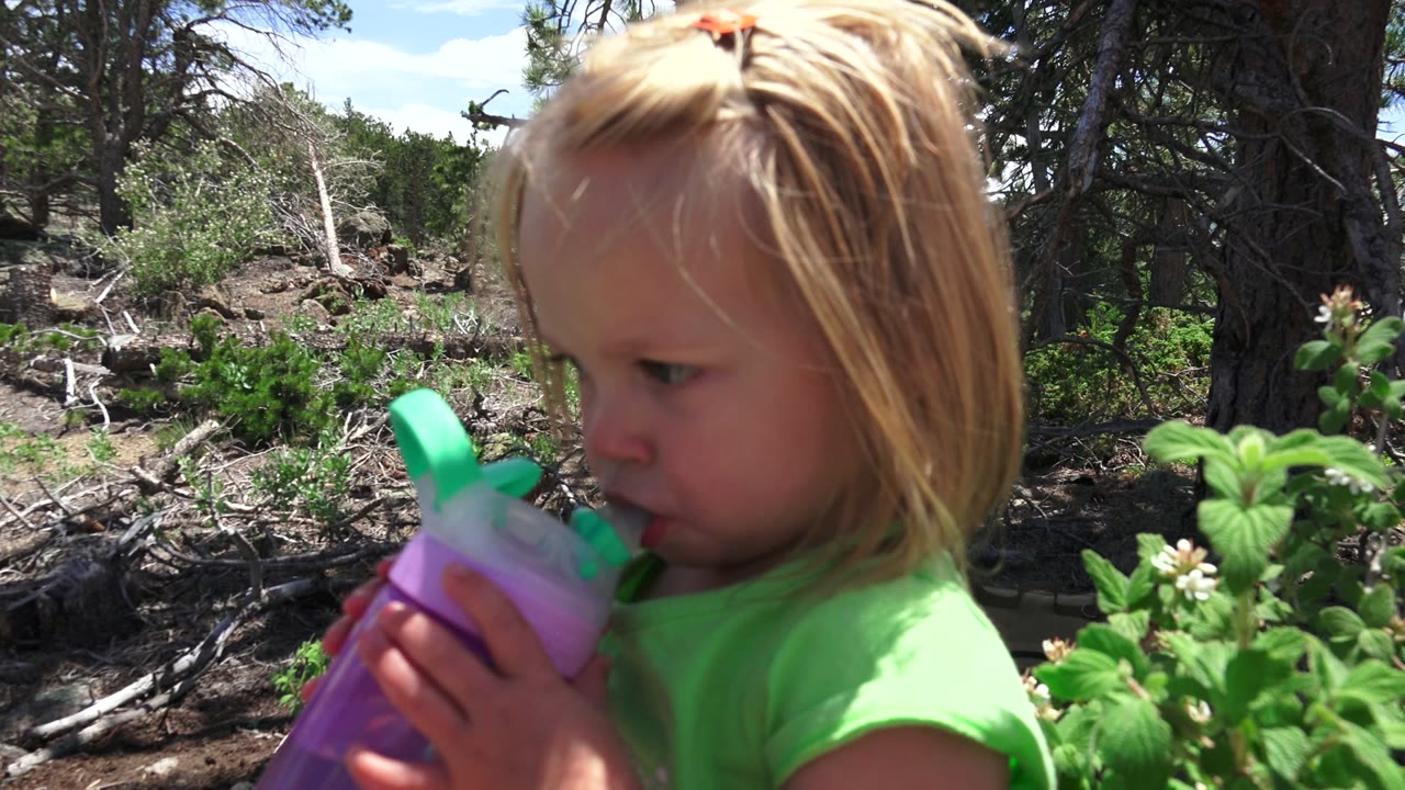Young blonde girl drinking from a purple cup beside green plants in a wooded area.