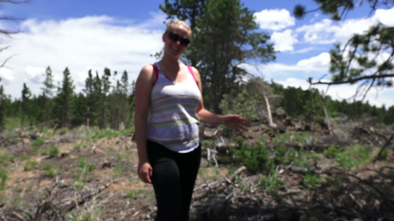 Woman in sunglasses stands outdoors near brush and trees.