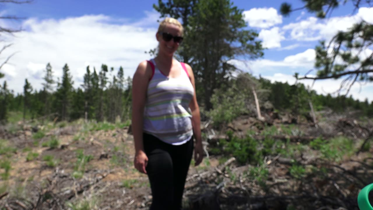 The woman shifts nearer the center of the Westminster, Colorado frame, standing in front of rough ground, low shrubs, and a line of evergreen trees.