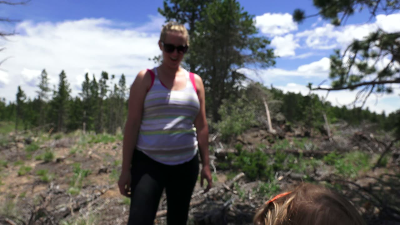 At 0:01 in Westminster, Colorado, the woman remains centered in the frame with a child's head low at the bottom edge, backed by small pines, rocky ground, and bright clouds.