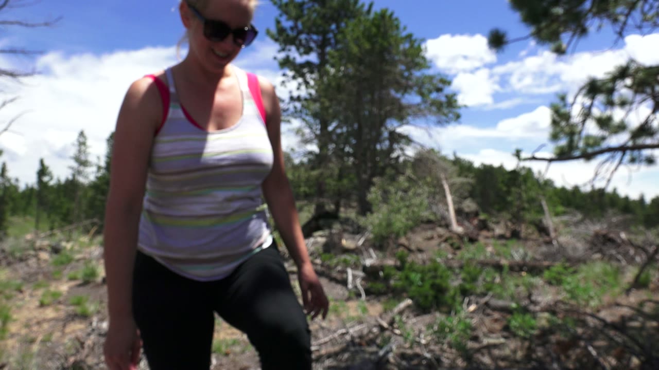 Woman in sunglasses steps closer in a wooded clearing.