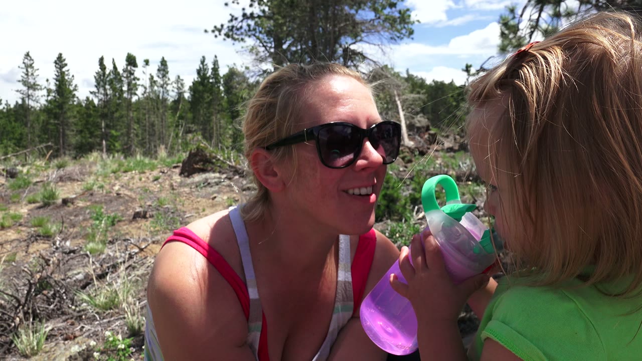 Mother in sunglasses smiling at a young girl holding a purple drink cup outdoors.
